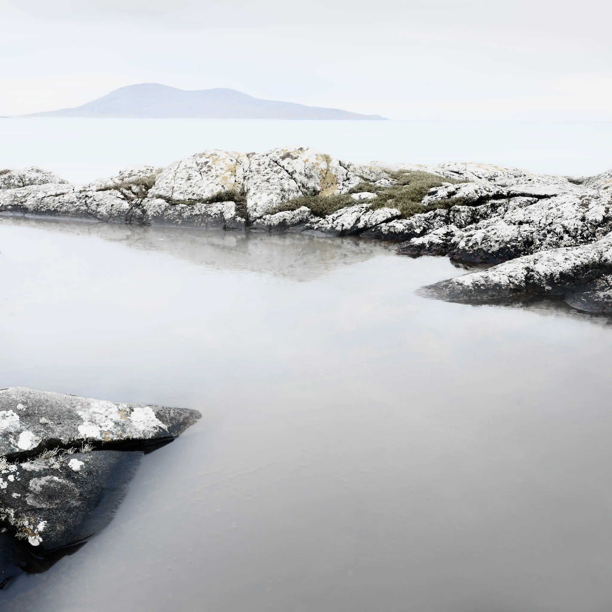 A rocky tidal pool ringed by lichen-covered rocks, an island silhouetted on a pale sea