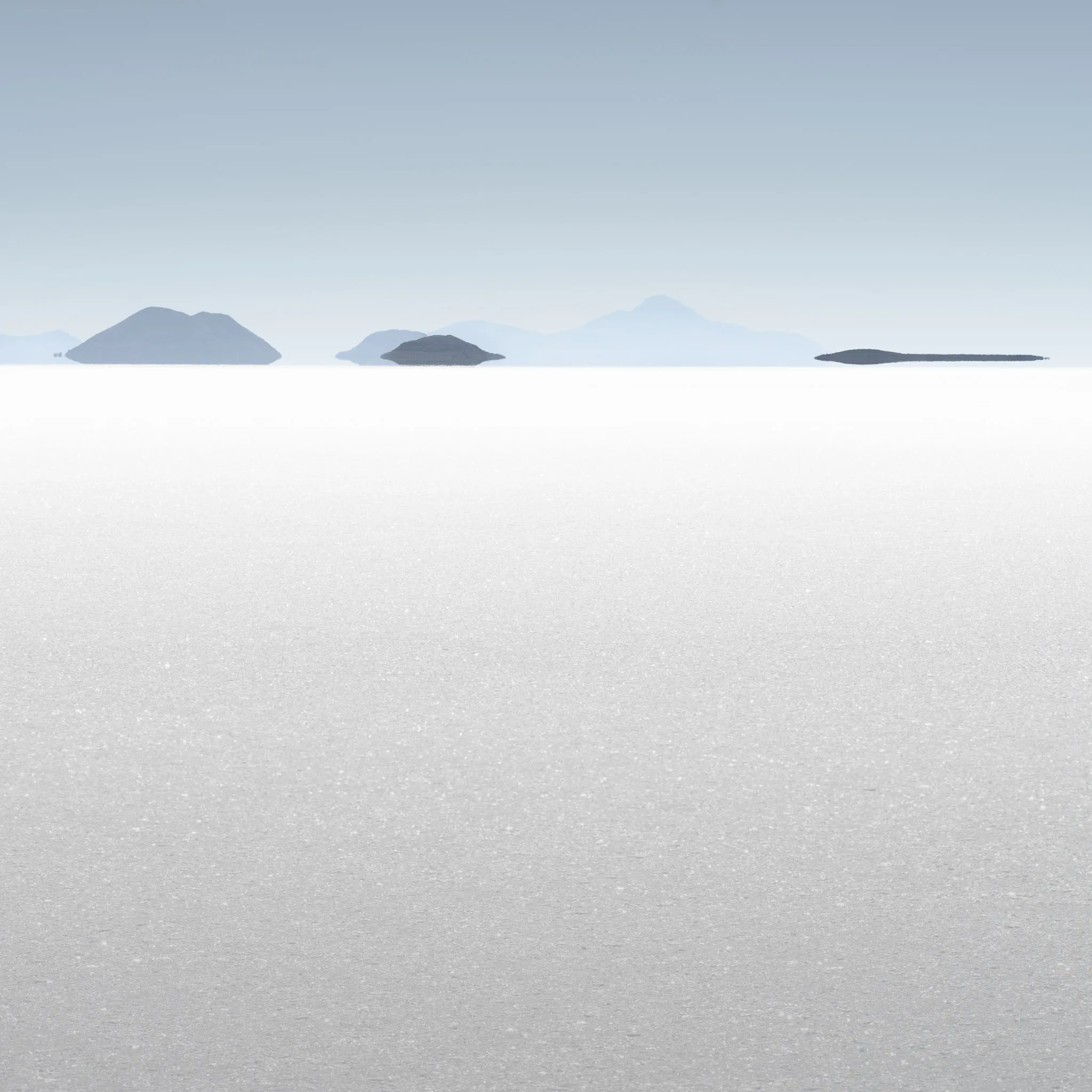 Distant islands and a faint volcano floating above the vast white expanse of a salt flat in high-key light