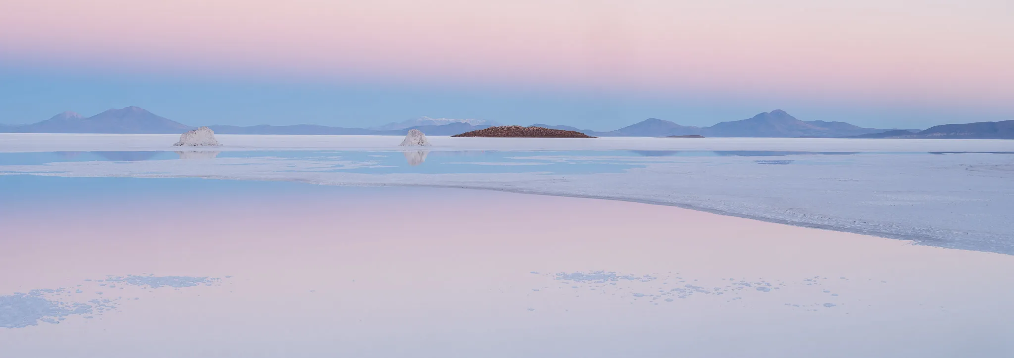 Salt cones and a low island reflected in a flooded salt flat under a soft pink twilight, distant volcanoes on the horizon