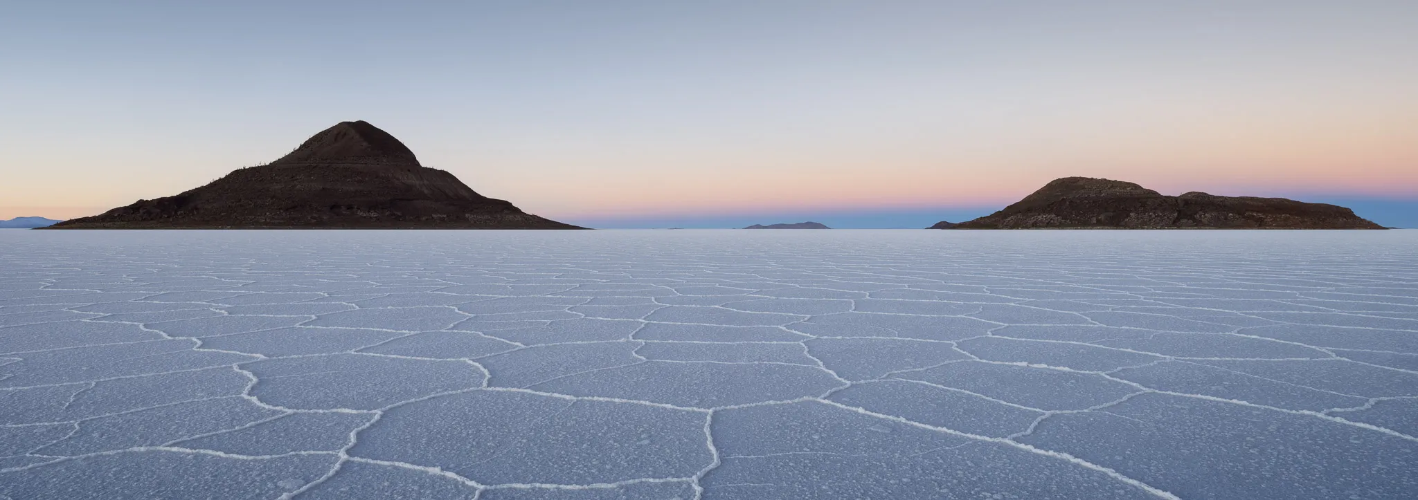 Two dark islands rising from the cracked salt flat at blue-and-gold twilight