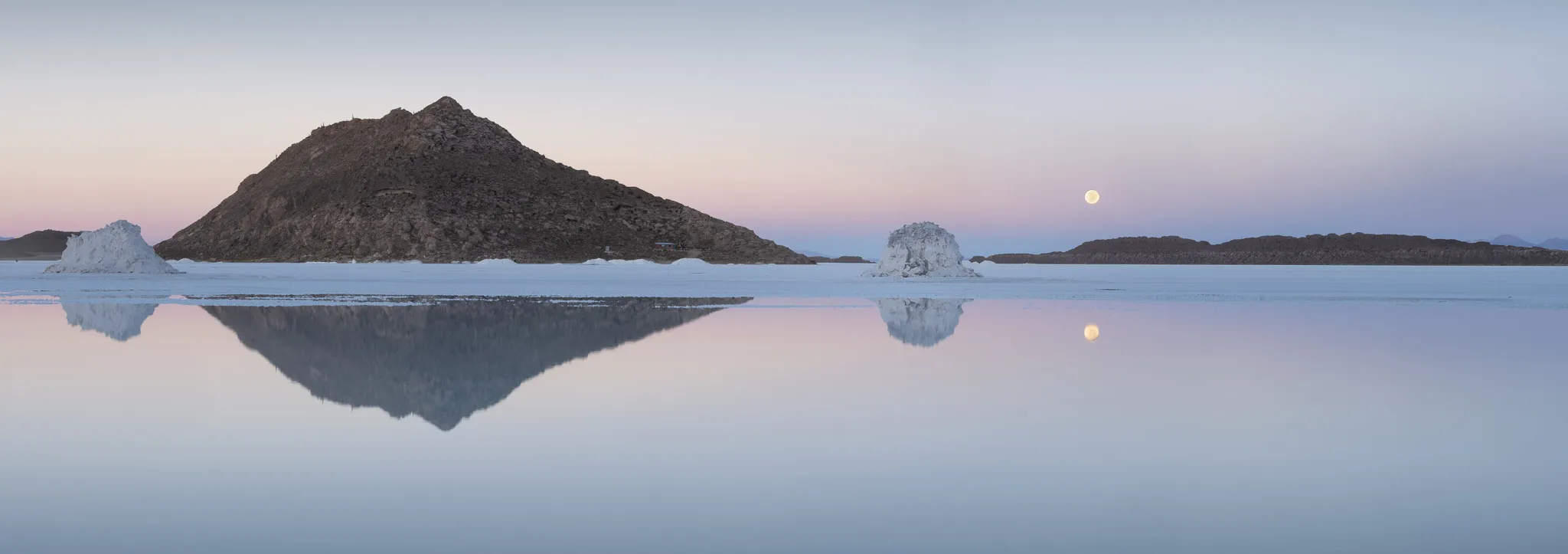 A rocky island and pale salt cones mirrored in still water as the moon rises in a pastel dusk sky
