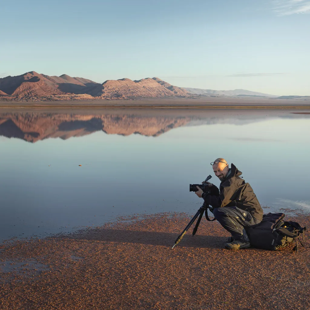 Thomas Senf crouching with his camera on a tripod at the edge of a still, mirror-like mountain lake at dawn